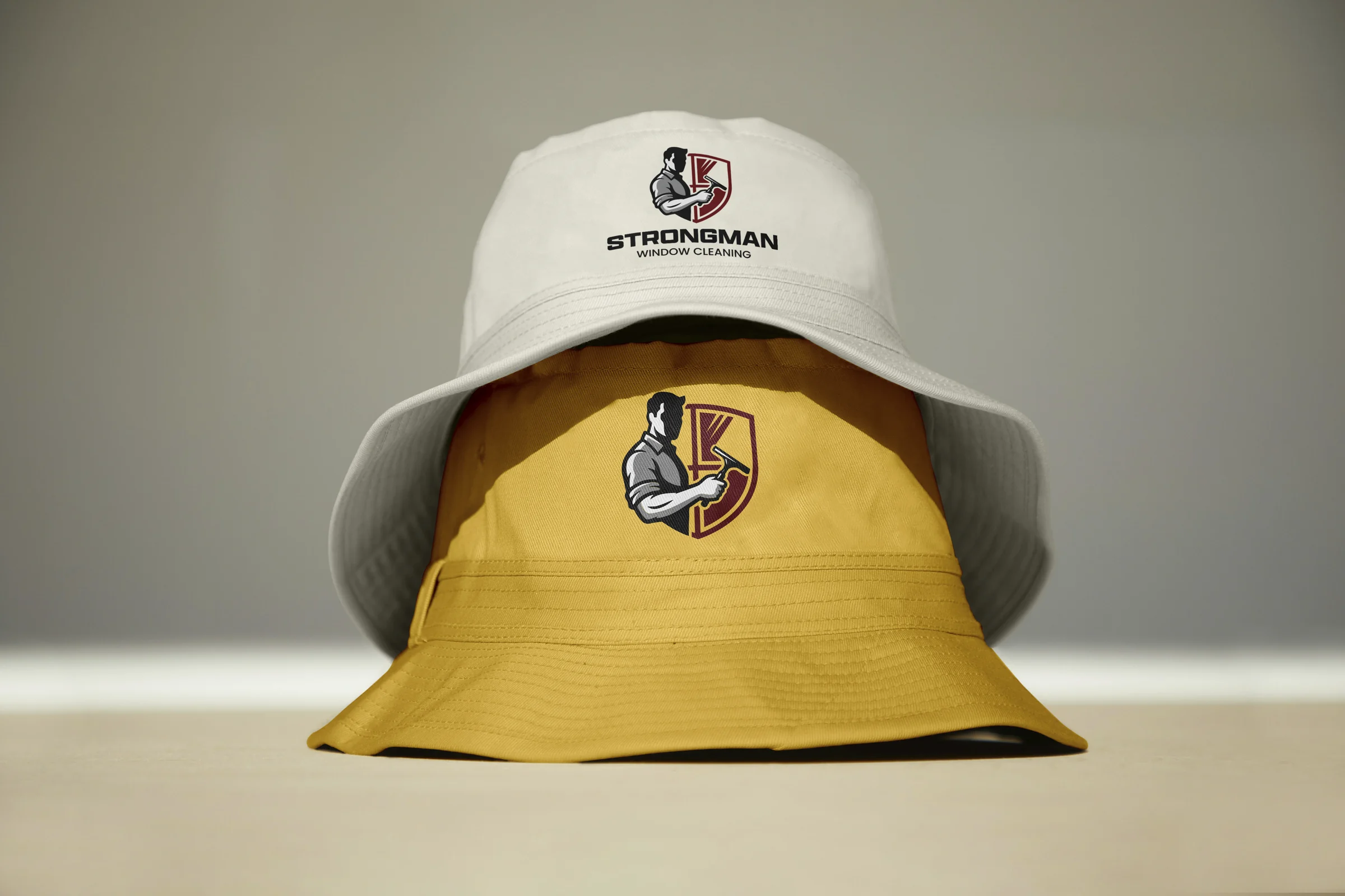 Two stacked Strongman bucket hats: a white hat with the full stacked logo on top of a gold hat with the shield mark alone